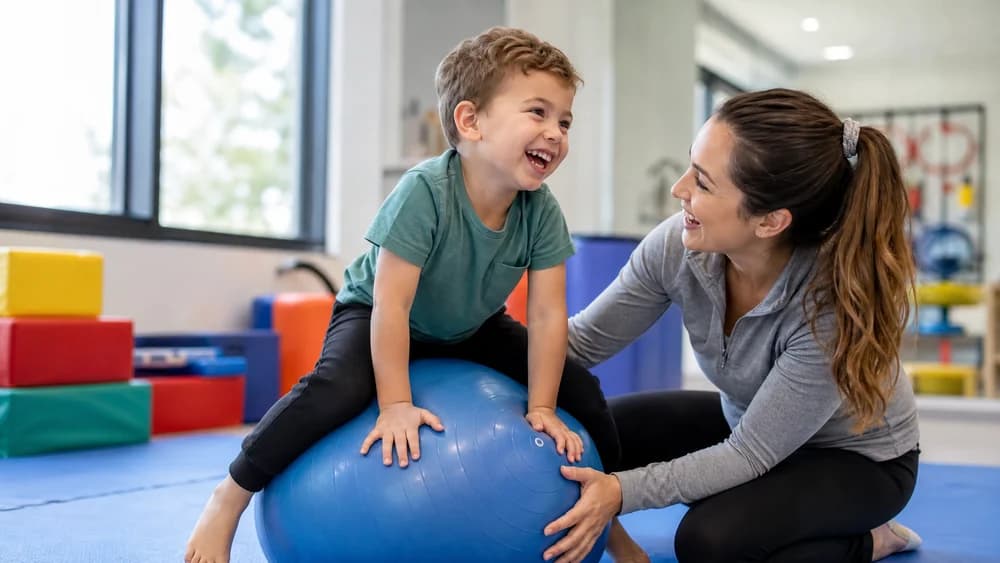 A laughing child balancing on a large therapy ball while a therapist steadies it and talks with him